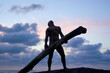 A naked man in silhouette lifts a heavy log on the beach as the spectacular sun sets behind him. 