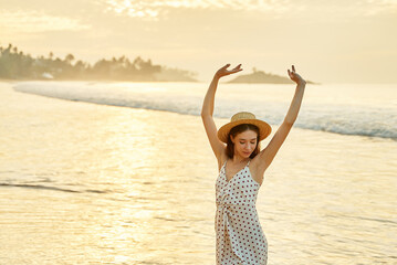 Woman in polka dot dress enjoys sunset at beach, arms raised high. Solo traveler celebrates freedom, warm summer evening by sea. Happiness, relaxation with ocean waves, sandy shore in background.