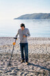 Young man with a metal detector walks along the beach, sweeping it over the pebbles