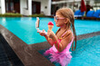 Little girl with sunglasses eats ice cream, streaming live on phone by poolside. Child in pink swimsuit enjoys summer treat engaging audience online. Pool activities, influencer lifestyle for kids.