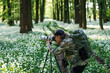 Landscape and wildlife photographer with backpack wearing camouflage shirt. Man with camera on tripod photographing nature in flowering spring woodland