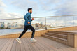 A bearded man in sportswear jogs on a waterfront boardwalk against an urban backdrop.