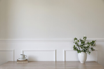 Wall Mural - Ceramic vase with blooming apple tree branches. Cup of coffee, tea on wooden table, desk with old books. Scandi home interior. Spring breakfast still life. Empty white wall mockup, stucco decor.