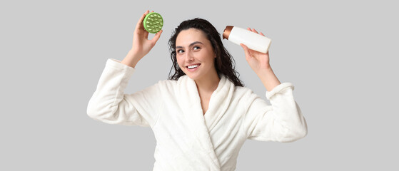 Happy young woman with hair scalp massager and bottle of shampoo on grey background