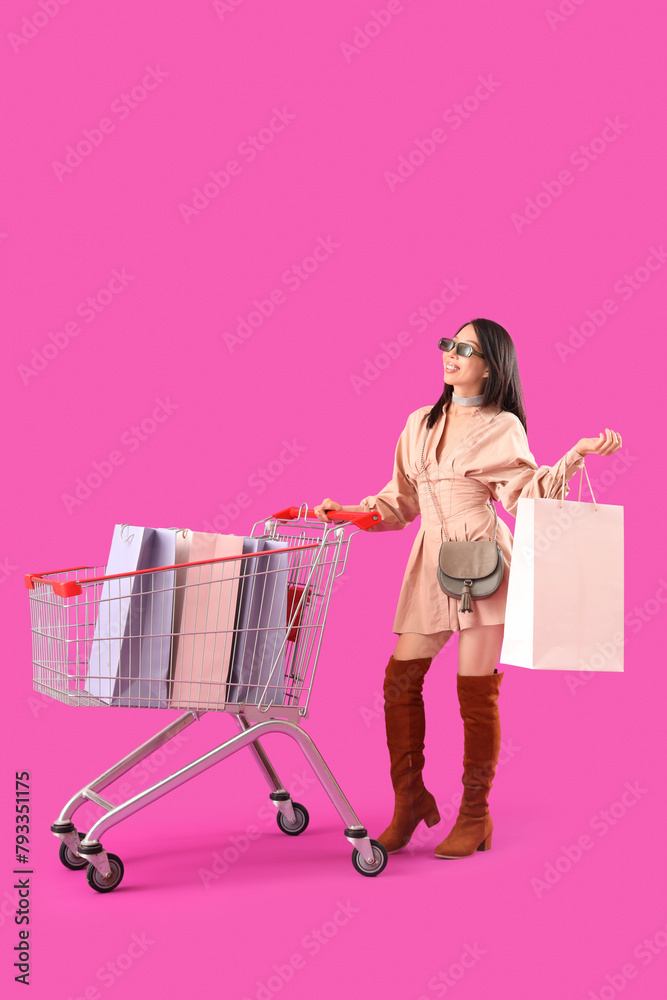 Beautiful young stylish Asian woman with trolley and shopping bags on purple background