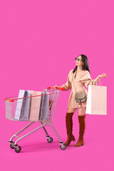 Sticker - Beautiful young stylish Asian woman with trolley and shopping bags on purple background