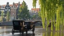 A Traditional Horse-drawn Carriage Crosses A Bridge Over A Tranquil Canal Lined With Historic Buildings And Lush Green Trees In Amsterdam, Netherlands. 