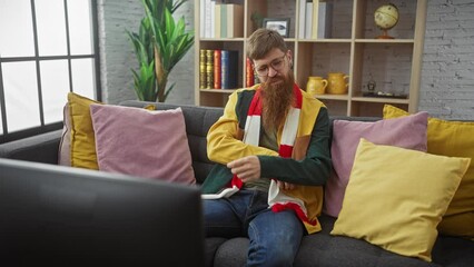 Wall Mural - A bearded man in glasses feels frustrated while sitting on a couch, wearing a colorful scarf, surrounded by colorful pillows.