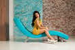 Lovely young woman make-up and long hair wearing yellow dress posing sitting on blue trendy comfortable sofa against white wall background. Fashion and interior design concept