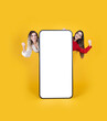 Two full body female  friends stand behind big huge blank screen mobile phone touch screen mock up do winner gesture clenched fist isolated over yellow studio background. Advertisement concept idea.