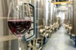 A close-up shot of a glass of red wine set against a blurred background of stainless steel wine fermentation tanks in a contemporary winery.