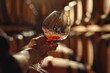 A close-up image of a hand holding a wine glass, elegantly swirling red wine, set against a blurred backdrop of a wooden wine cellar filled with barrels.