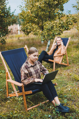 Wall Mural - Concentrated woman in the garden working on a laptop while sitting on a chair among the trees. A woman works remotely from the garden, thinking about a new business or creative idea.