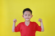 Smiling Asian little boy showing his muscle with looking camera isolated on yellow background. Kid show strong gesture.