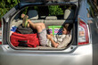 Cute little boy laying on the back of the bags and baggage in the car trunk ready to go on vacation with happy expression. Kid resting playing on smartphone