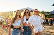 Three smiling young women walk hand in hand, sunny beach music festival. Friends in summer outfits with sunglasses, dance, socialize in coastal event. Youth culture, music on sandy shore.