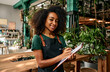 Management of flower shop. Pleasant black woman engaging in inventory work in floral boutique while standing and smiling at camera. Charming lady in green apron noting all details on a clipboard.