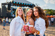 © artiemedvedev - Girls cheer colorful drinks at beach music fest. Happy friends enjoy sunny day, summer party vibes. Dance, laughter near sea with crowd, stage in background. Youth celebrating, toasting at festival.