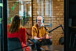 © .shock - A gathering of young business professionals, some seated in a glass-walled office, engage in a lively conversation and record an online podcast, embodying modern collaboration and dynamic interaction