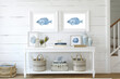 Recibidor con decoración marinera con pared de madera de tablones blancos, dos cuadros simétricos con peces de color azul y consola de madera blanca con decoraciones 