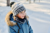 Fototapeta  - Cute smiling boy in grey knitted beanie hat and blue winter jacket looking at camera while walking along road in park or forest