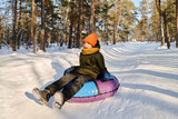 Fototapeta  - Happy cute boy in warm winter jacket, beanie and scarf sitting on snow tube while moving down hill in natural environment