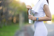 Attractive young Asian woman who is thirsty is drinking water while taking a break from jogging at the park. Asian woman uses a towel to wipe away her sweat while taking a walk to rest from jogging.