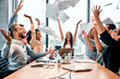 Celebrating success. Joyful command throwing up documents in air above long wooden table with excited expression. Playful business people reaching goal and entertaining together as sign of victory.