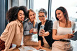 Team of different successful beautiful happy business women at work in the office. Sincere lady in the work team stand and have a friendly conversation with a cup of coffee and look at the phone.