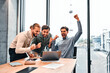 Three adult business men in formal clothes celebrate with gestures and emotions a successful online deal with partners while sitting in an office with panoramic windows and a view of the city.