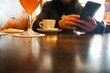Frau sitzt mit Handy in Händen vor braunem Holztisch mit Cocktailglas und weißer Tasse Kaffee in Restaurant am Mittag im Frühling