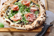 Wood fired Italian pizza with prosciutto, parma ham, arugula, and parmesan, on a wooden cutting board, top view.