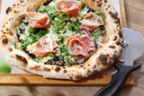 Fototapeta Tulipany - Wood fired Italian pizza with prosciutto, parma ham, arugula, and parmesan, on a wooden cutting board, top view.