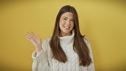 Canvas Print - Joyful young hispanic woman gives warm welcome with hello gesture, wearing sweater, standing over yellow isolated background, beaming a confident smile while waving hand