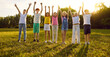 © Studio Romantic - Group of a smiling kids friends raising hands up on green grass in the park standing in a line. Happy children having fun together outdoors on a sunny summer day in casual clothes in the camp.