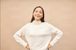 Studio portrait of pretty confident asian girl in mockup beige sweater standing with satisfied smile and holding hands on waist after fulfilling task or achieving goal, motivating you to join her