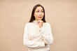 Portrait of beautiful asian woman standing against pink background in beige turtleneck, touching her chin and looking upwards as if hatching an idea or contemplating decision or plan