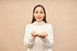 Studio portrait of asian girl in sweater standing with empty palms together like holding something in hands with generosity making receiving or giving gesture, isolated against pink background