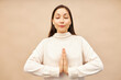 Pretty gentle woman meditating against pink studio background in beige sweater, standing with closed eyes, doing namaste gesture with hands, reaching harmony. Yoga, meditation and zen concept