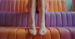 Close-up of a woman's legs sitting on a soft bleacher. Woman wearing socks sitting on a purple bleacher on neutral background.