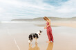 Caucasian blonde woman in a summer dress with her border collie dog taking a walk on the beach