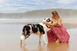 young caucasian woman crouching on the sand of the beach petting and cuddling her border collie dog during a summer stroll