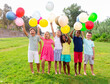 Portrait of cheerful emotional preteen boys and girls of different nationalities with colorful toy balloons in hands posing on green lawn in city park 