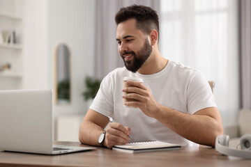 Canvas Print - Young man with cup of coffee watching webinar at table in room