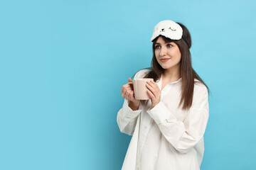 Wall Mural - Woman in pyjama and sleep mask holding cup of drink on light blue background, space for text