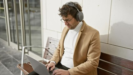 Wall Mural - Handsome hispanic man with headphones using laptop on city bench outdoors