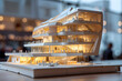 A model of a modern building with white and light wood features. It has several stories and a curved design. The model is displayed on a white surface and there are miniature people placed on the buil
