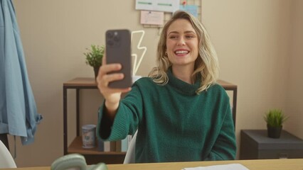 Poster - A smiling young caucasian woman in a green sweater takes a selfie indoors with her smartphone.