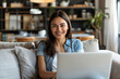 © Emanuel - Attractive, smiling woman of ethnic background engaged in remote work from her residence, utilizing a laptop computer