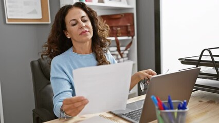 Sticker - Mature hispanic woman reviewing documents at a laptop in a modern office interior.